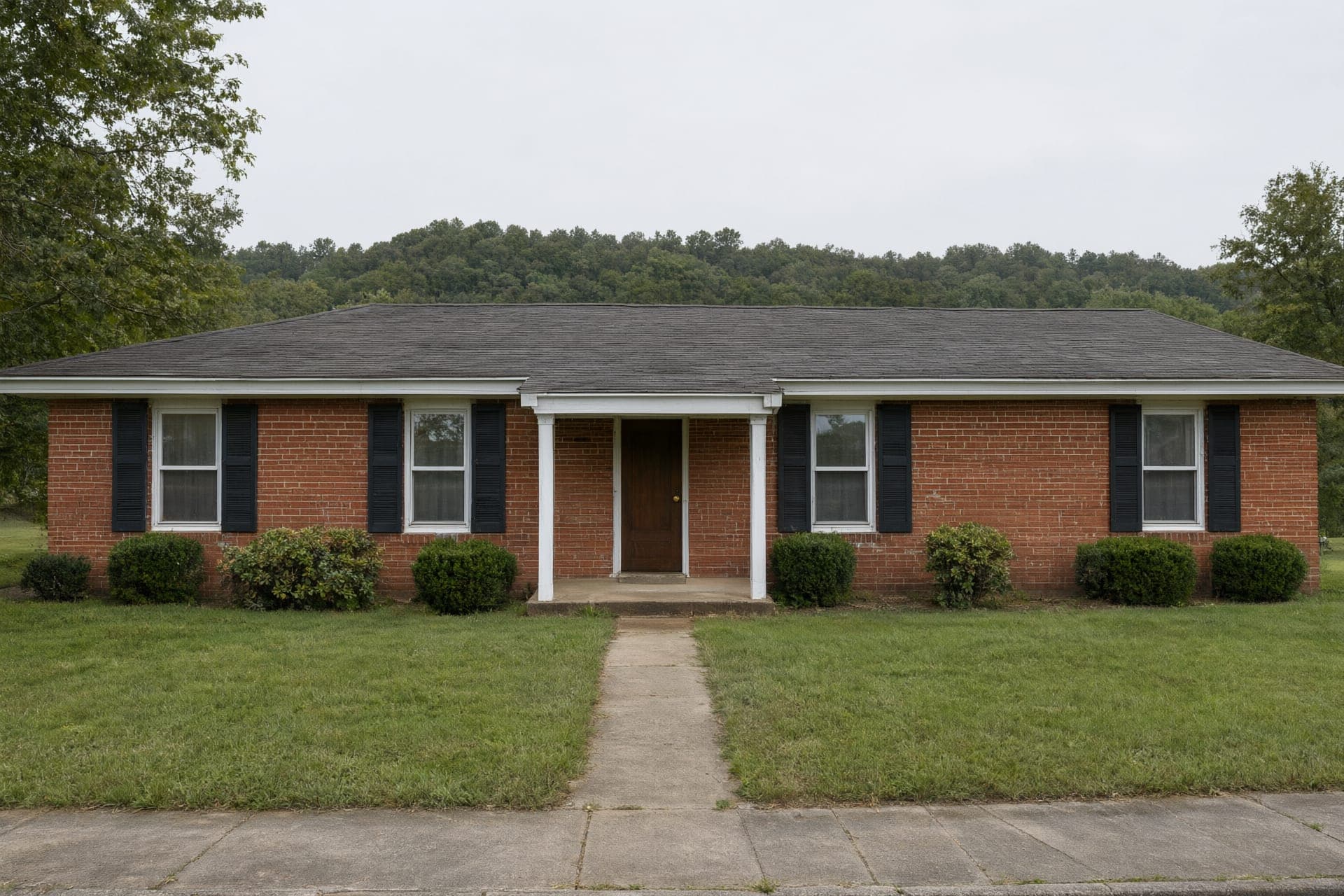 1970s single-story red brick ranch house with black shutters and a small front porch, wooded ridge behind