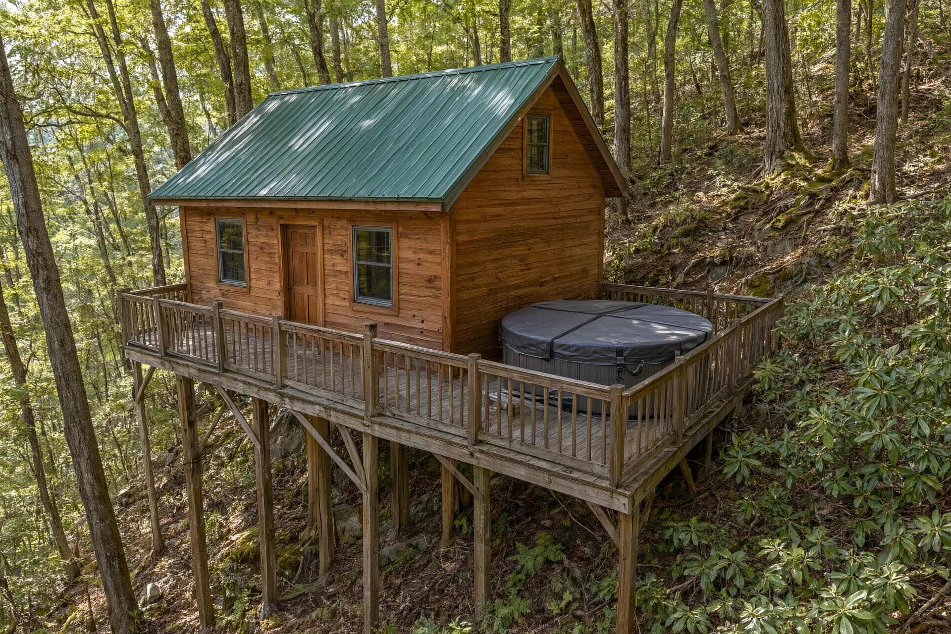 Wooden rental cabin with a green metal roof and a covered hot tub on a deck held up by tall posts on a steep slope