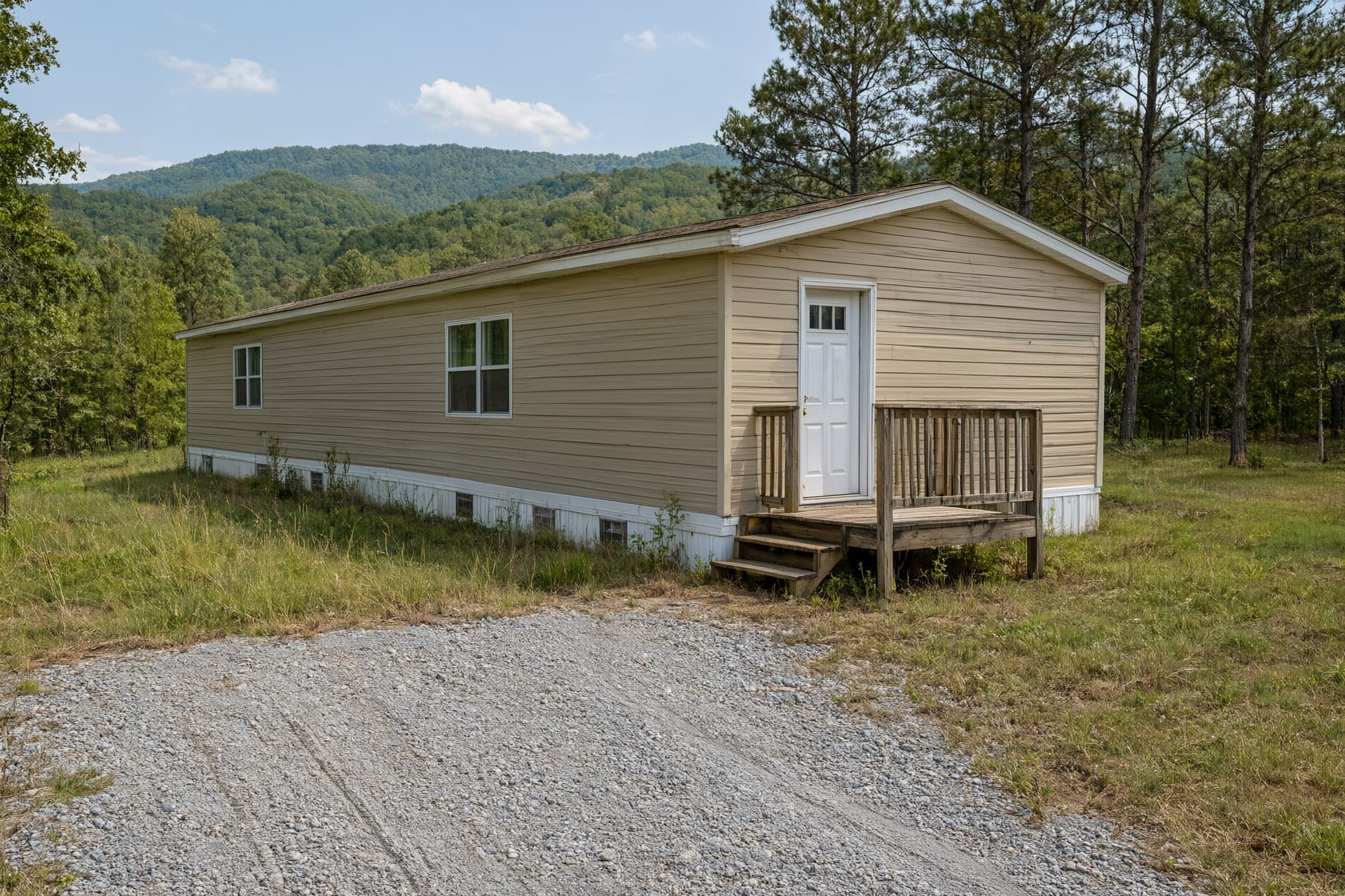 Tan manufactured home with white skirting and a wooden front deck on a rural lot with hills behind